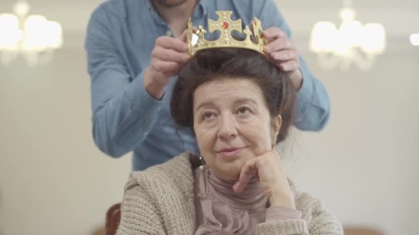 Porträt einer hübschen, reifen Frau. Der erwachsene Enkel bringt die Krone und setzt sie der Oma auf den Kopf, die Seniorin ist überrascht, sie ist glücklich und lächelt. Familienbeziehung — Stockvideo