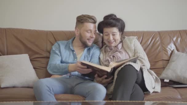 Schattige mooie oma en volwassen kleinzoon zittend thuis op de bruin lederen sofa kijken naar oude Foto's in het grote fotoalbum. Familie Onthoud het verleden, herinner oude tijden, deel herinneringen — Stockvideo