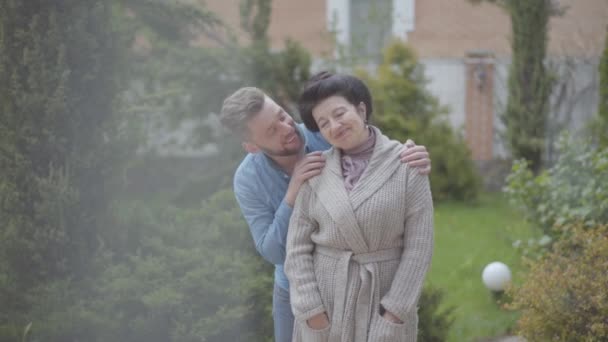 Портрет щасливої дорослої жінки, що стоїть в саду перед великим будинком, дорослий онук обіймає її, поклавши руку на плече. Ніжні стосунки в сім'ї, кохання, підтримка — стокове відео