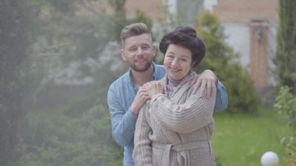 Portrétní krásná dospělá žena stojící v zahradě před velkým domem, dospělý vnuk, který ji objal a položil jí ruku na rameno. Něžný vztah v rodině, lásce, podpoře — Stock video