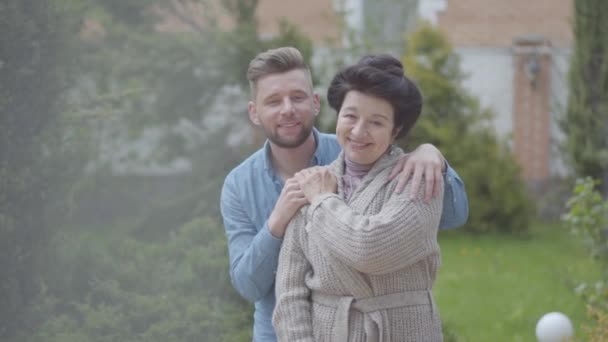 Retrato linda mujer madura de pie en el jardín en frente de la casa grande, nieto adulto abrazándola, poniendo la mano sobre su hombro. Relación tierna en familia, amor, apoyo — Vídeo de stock