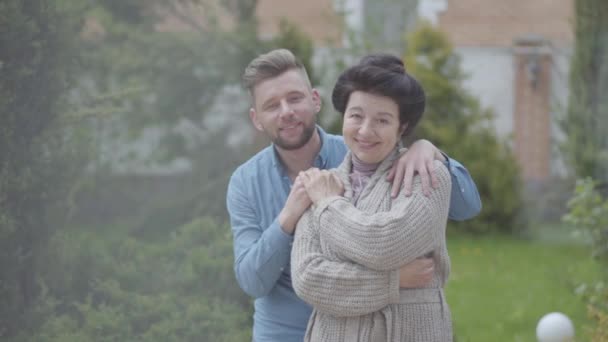 Ευτυχισμένη ώριμη γυναίκα στέκεται στον κήπο μπροστά από το μεγάλο σπίτι, ενήλικο εγγονό της αγκαλιάζοντας την, βάζοντας το χέρι στον ώμο της. Τρυφερή σχέση στην οικογένεια, την αγάπη, την υποστήριξη — Αρχείο Βίντεο