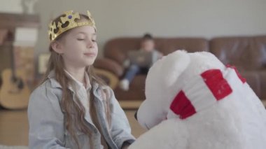 Sevimli kız bir oyuncak kuzey ayısı ile oynarken, onu bir taç ve onun ikiz kardeşi kanepede dizüstü bilgisayar oynarken soyunma