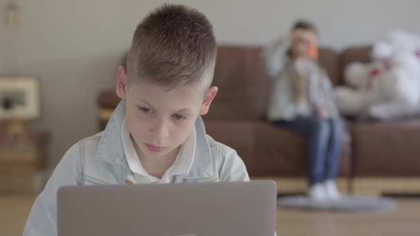 Piccolo ragazzo seduto sul computer portatile e piccola ragazza, la sua sorella gemella soffia bolle di sapone sul divano di casa — Video Stock