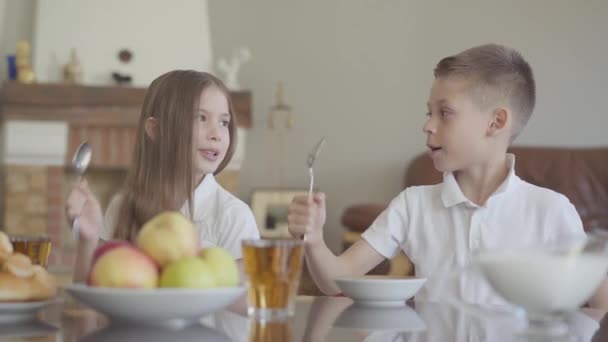 Bliźniacy brat i siostra siedzą przy stole w białych koszulkach przed szkołą i pukają z łyżkami, krzycząc z radością na jedzenie — Wideo stockowe