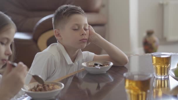 学校の前に欲望なしで朝食のためにお粥やコーンフレークを食べようとしている姉妹と彼女の疲れた兄弟の双子の肖像画. — ストック動画