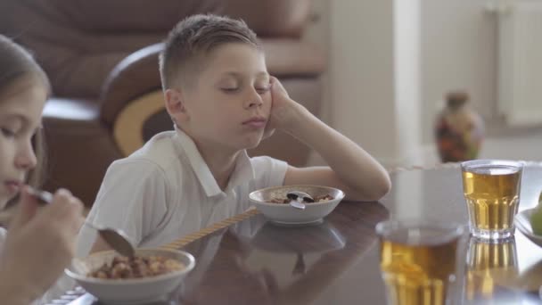 Portret van zuster en haar vermoeide broer tweeling proberen te eten pap of cornflakes voor ontbijt zonder verlangen voor school. — Stockvideo