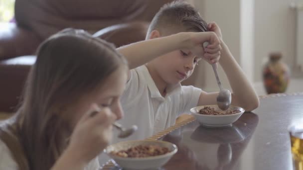 Portret schattig zus en haar vermoeide broer tweeling proberen te eten pap of cornflakes voor ontbijt zonder verlangen voor school. — Stockvideo