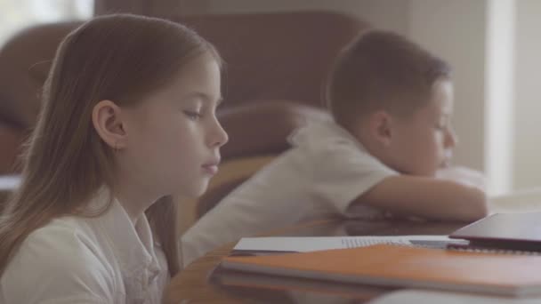 Syster och bror sitter på bordet och gör läxor från skolan. Flickan somnar på bordet. Pojke läser fortfarande en bok. — Stockvideo