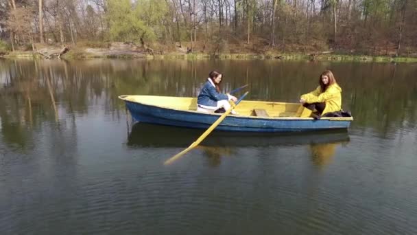 Zwei junge hübsche Mädchen, die in dem kleinen Boot inmitten eines wunderschönen spiegelnden Sees oder Flusses sitzen. aktiver Lebensstil, Verbindung mit der Natur. Seitenansicht — Stockvideo