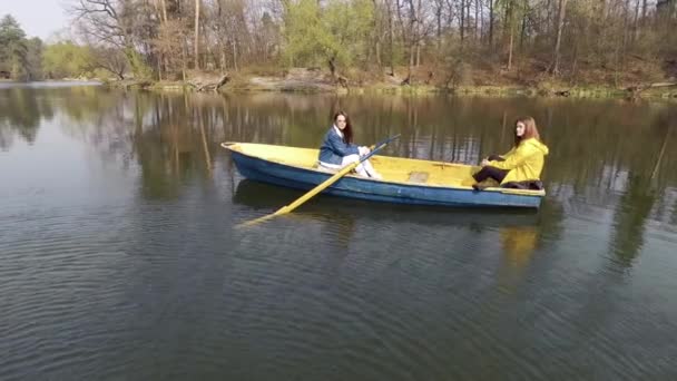 Dos chicas guapas jóvenes sentadas en el pequeño bote en medio de un hermoso lago o río reflectante. Estilo de vida activo, conexión con la naturaleza. Vista lateral — Vídeos de Stock