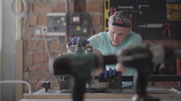 El hombre con gafas de protección cortando una tabla de madera con una pequeña sierra circular en la mesa en el taller. Fábrica de muebles. Concepto de fabricación manual. Artesano trabaja en un taller . — Vídeo de stock