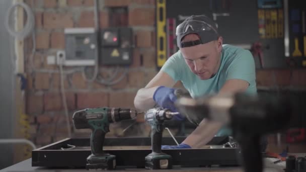 Snickaren i svart mössa och handskar sätta lim på träram. Mannen som använder silikon limpistol i möbeltillverkning. Joiner gör sitt arbete i fabriken. Screwdrives på den. — Stockvideo