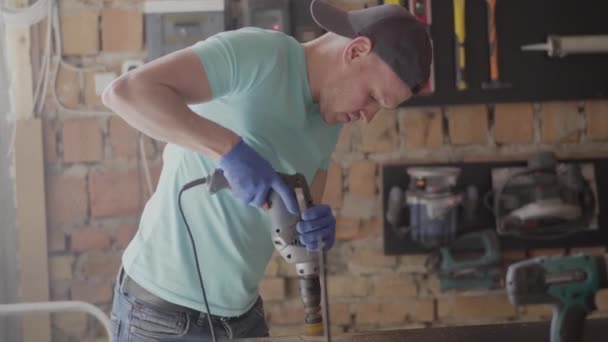 Retrato de joven ingeniero maestro enfocado en perforar un agujero con herramienta en el fondo de un pequeño taller. Concepto de fabricación manual. Artesano trabaja en un taller . — Vídeo de stock