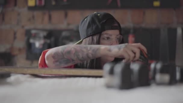 Young woman in a cap and protective glasses with pierced lip and ring in nose measuring length of the MDF plate with the measuring tape on the factory. Beautiful unusual woman, carpenter profession — 비디오