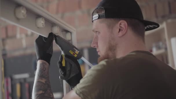 El carpintero en gorra negra y guantes con tatuajes en los brazos poniendo pegamento en el marco de madera para el espejo. El trabajador que utiliza pistola de pegamento de silicona en la fabricación de muebles. Artesano trabaja en un taller . — Vídeos de Stock