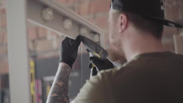 De timmerman in zwarte dop en handschoenen met tatoeages op armen zetten lijm op het houten frame voor de spiegel. De werknemer met behulp van silicone lijm pistool in meubelproductie. Craftman werkt in een workshop. — Stockvideo