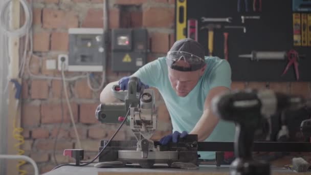 Retrato habilidade artesão em óculos de proteção cortando uma tábua de madeira com uma pequena serra circular sobre a mesa na oficina. Fábrica de móveis. Conceito de fabricação manual. Artesanato trabalha em um — Vídeo de Stock