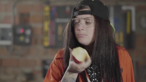 Portret vaardigheid craftswoman het eten van de appel op de voorgrond close-up. De man die met het hout op de achtergrond werkt. De vrouw heeft een pauze op het werk. Werkproces in de meubelfabriek. — Stockvideo