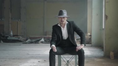 Bir fedora şapka ve klasik bir takım elbise ile destoyed garajda sandalyede oturup adam ve bir yazı tura. Terk edilmiş harap bina.