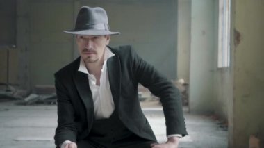 Portre kendine güvenen adam bir fedora şapka ve klasik bir takım elbise ile destoyed garajda sandalyede oturur ve bir yazı tura. Terk edilmiş harap bina.