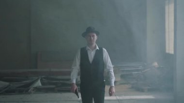 Terk edilmiş bir binada elinde silahla kameraya yürüyen şapkalı yakışıklı kendine güvenen bir adam portre. Mafyanın başı boş bir binada. Havalı adam, haydut, mafya. Yavaş çekim.