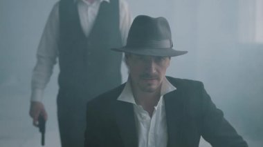 Şapkalı yakışıklı, terk edilmiş bir binada kameraya bakıyor. Arkadan gelen ve silahı adamın kafasına dayayan tanınmayan bir adam. Boş bir binada mafya. Yavaş çekim.