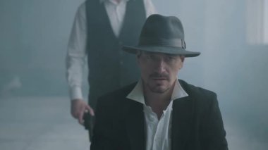 Portre emin kötü adam fedora şapka ve diğer adam bir silahla arkasından yürüyen sandalyeye oturup ve kafasına ateş. Arkadan gelen ve silahı mans kafasına dayayan tanınmayan adam.