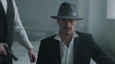 Portre kendine güvenen adam fedora şapka ve diğer adam bir silahla arkasından yürüyen sandalyeye oturup ve kafasından onu vur. Arkadan gelen ve silahı mans kafasına dayayan tanınmayan adam.