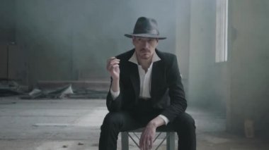 Portre kendine güvenen adam bir fedora şapka ve klasik bir takım elbise ile destoyed garajda sandalyede oturur ve bir yazı tura. Terk edilmiş harap bina. Terk edilmiş harap bina.