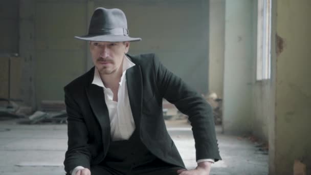 Porträt eines selbstbewussten Mannes, der mit Fedora-Hut und klassischem Anzug auf dem Stuhl in der verlassenen Garage sitzt und eine Münze wirft. Verlassenes, marodes Gebäude. — Stockvideo