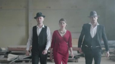 Şapkalı ve takım elbiseli iki kendine güvenen adam ve terk edilmiş bir binada kameraya doğru yürüyen kırmızı elbiseli kadın. Boş bir binada mafya. Havalı adamlar, haydut, mafya, suç çetesi