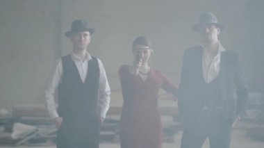 Terk edilmiş bir binada kameraya tabanca doğrultan kırmızı elbiseli bir kadının iki yanında duran iki kendinden emin iyi giyimli adam. Havalı adamlar, haydut, mafya, suç çetesi