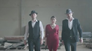 Terk edilmiş bir binada kameraya tabanca doğrultan kırmızı elbiseli bir kadının iki yanında duran kendine güvenen iki iyi giyimli adamı portre. Havalı adamlar, haydut, mafya, suç çetesi