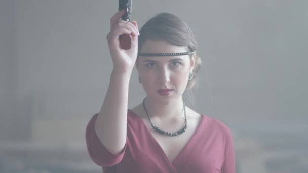 Confident attractive woman in stylish red dress is aiming a pistol at the camera in an abandoned dusty building, then lowers the gun. Dangerous lady. Retro, vintage style — Stock Video