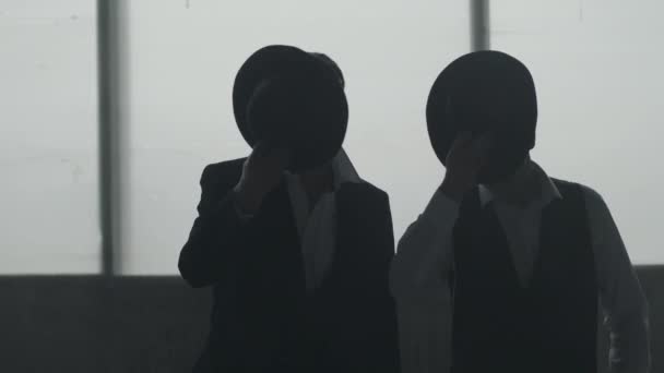 Twee donkere figuren van zelfverzekerde goed geklede mannen in hoeden die in de buurt van elkaar staan voor groot raam. Tegelijkertijd zetten beide mensen hoeden op hoofden. Straf autoriteit, maffia — Stockvideo