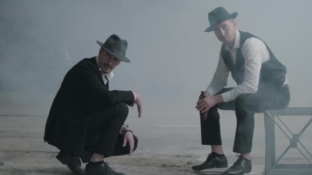 Two stylish men in hats, one sitting on the chair, another squatting in an abandoned building. The mafia guys have meeting in an empty building. Cool guys, thugs, mafia, criminal gang — Stock Video