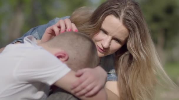 The little boy sitting on the grass hiding his face, a young beautiful woman caressing his head, trying to cheer him up. A mother comforts her son. A loving family, support in a difficult situation — Stock Video