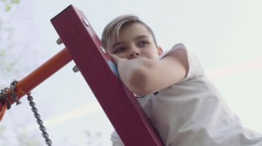Sarışın çocuk bir salıncak tırmandı ve yukarıdan aşağıya kameraya bakar. Aktif yaşam tarzı, kaygısız çocukluk, sevimli çocuk açık havada oynarken