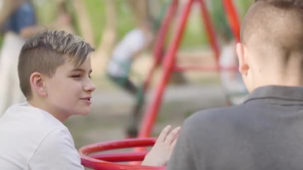 Два брата сидят в парке и разговаривают. Женщина раскачивает мальчиков на заднем плане — стоковое видео