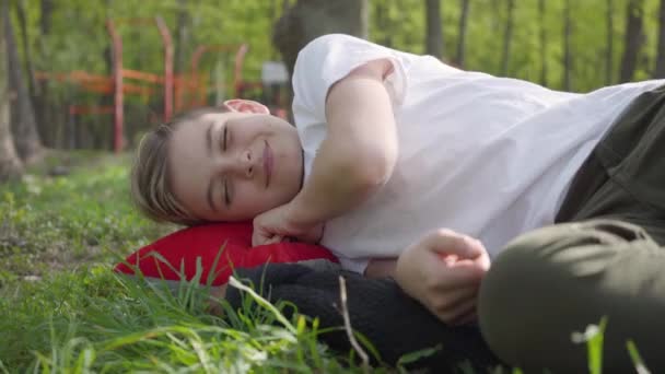 Junge liegt an einem warmen Frühlingstag im Park in der Nähe des Spielplatzes aufgeregt oder müde nach der Schule auf dem Rasen — Stockvideo
