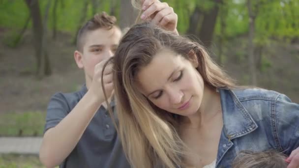 Retrato hijo haciendo un peinado a su madre o niñera mientras están en el parque en soleado día de primavera y hablando. recreación al aire libre . — Vídeo de stock