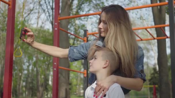 Madre con el pelo largo rubio haciendo una foto con sus dos hijos pequeños en el patio de recreo — Vídeos de Stock