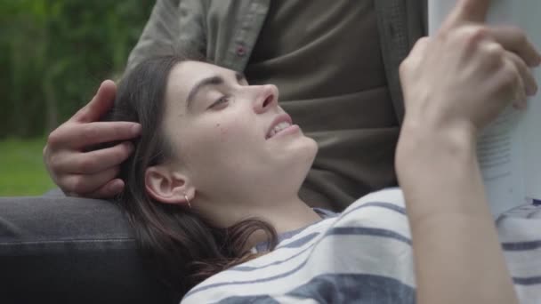 Close up retrato jovem casal em roupas casuais passar tempo juntos ao ar livre, tendo data. A menina bonita deitada às voltas do namorado, lendo o diário de perto. Lazer de verão — Vídeo de Stock