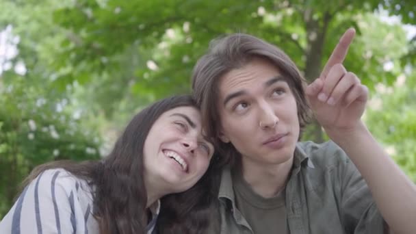 Portrait d'un jeune couple souriant en vêtements décontractés passant du temps ensemble dans le parc, ayant un rendez-vous. Le gars pointant vers le haut avec son doigt tandis que la dame s'appuie sur son épaule — Video