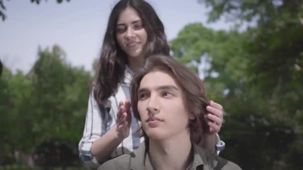 El joven guapo con aparatos ortopédicos y pelo largo sentado en primer plano, su novia viene por detrás y cubre sus ojos. El tipo la mira. Preciosa pareja tiene una cita en el parque — Vídeo de stock