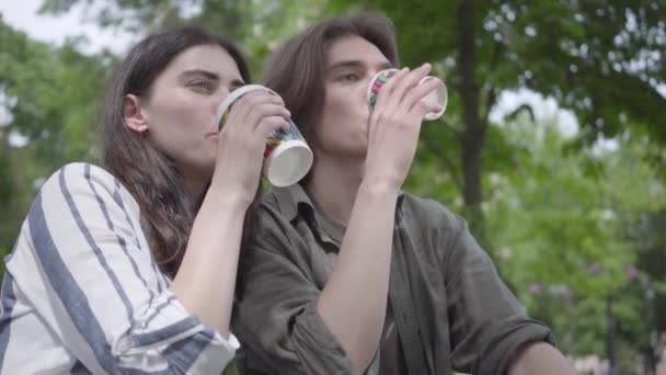 Jovem casal feliz em roupas casuais passar o tempo juntos no parque, tendo um encontro. Os alunos bebendo café, menina compartilhando sua bebida com o namorado, mas ele não gosta do sabor — Vídeo de Stock