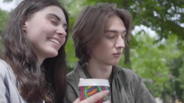 Retrato jovem casal feliz em roupas casuais passar o tempo juntos no parque, tendo um encontro. Os alunos bebendo café, menina compartilhando sua bebida com o namorado, mas ele não gosta do sabor — Vídeo de Stock