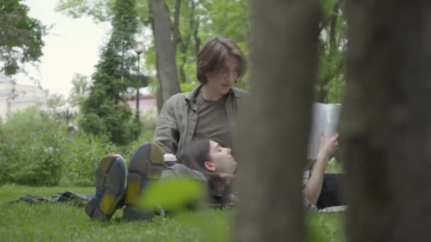 Porträtt ungt par spenderar tid tillsammans utomhus, har datum. Killen sitter på täcket på gräset, vacker flicka som ligger på hans varv läsa tidningen. Sommar fritid — Stockvideo