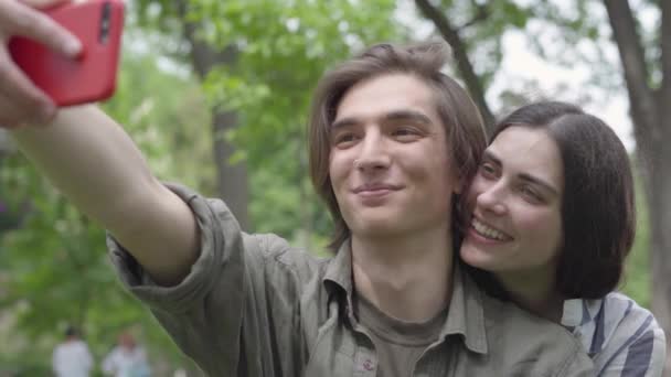 Retrato lindo par de estudiantes tomando selfie sentado en el césped en el parque. El tipo está en primer plano, la chica lo abraza por detrás. Tiempo libre de verano. Adicción a los aparatos — Vídeos de Stock